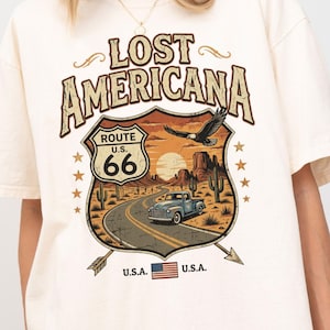 May include: Cream-colored t-shirt featuring a graphic design with the text "LOST AMERICANA" and "ROUTE 66". The design includes a road, car, desert landscape, and an eagle. The shirt has short sleeves and a relaxed fit.