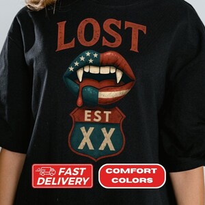 Puede incluir: Camiseta negra con un diseño gráfico que presenta la palabra "LOST" en rojo, un par de labios de vampiro con una combinación de colores patrióticos y una señal de tráfico que dice "EST XX". La camiseta también tiene dos rectángulos rojos y blancos con texto.