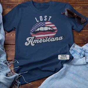 May include: Navy blue t-shirt with the words "LOST Americana" above a graphic of lips designed with the American flag. The shirt is paired with sunglasses and light blue denim jeans on a wooden surface.
