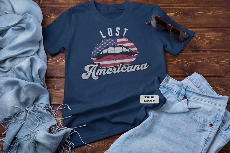 May include: Navy blue t-shirt with the words "LOST Americana" above a graphic of lips designed with the American flag. The shirt is paired with sunglasses and light blue denim jeans on a wooden surface.