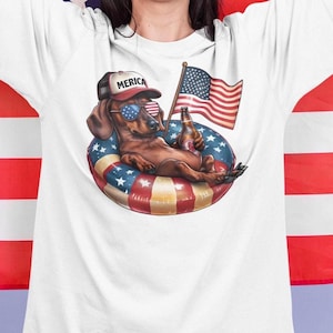 Pode incluir: Camiseta branca com um dachshund usando óculos de sol e um chapéu com a palavra "MERICA". O cão está relaxando em um anel inflável com um design de bandeira americana, segurando uma garrafa de cerveja e acenando com uma bandeira americana.