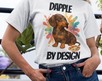 May include: White t-shirt featuring a dachshund illustration with a colorful paint splash background. The text "DAPPLE BY DESIGN" is printed above and below the dog image. The shirt is made of a soft material.