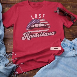 May include: A red t-shirt with the words "LOST Americana" and a graphic of lips with an American flag design. Sunglasses rest on the shirt, and blue jeans are folded nearby.