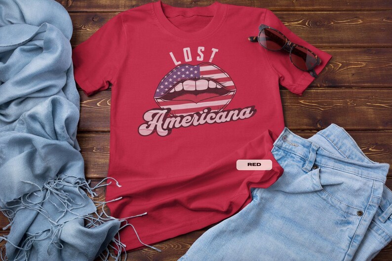 May include: A red t-shirt with the words "LOST Americana" and a graphic of lips with an American flag design. Sunglasses rest on the shirt, and blue jeans are folded nearby.