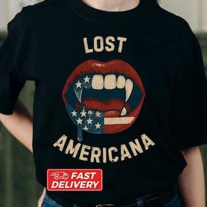 Puede incluir: Camiseta negra con un gráfico de labios rojos con colmillos y un diseño de la bandera estadounidense. El texto "LOST AMERICANA" está encima de los labios, con la palabra "AMERICANA" debajo. Una pegatina roja "FAST DELIVERY" está en la parte inferior.