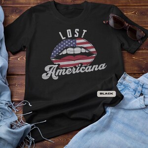 May include: Black t-shirt with the text "LOST Americana" above a graphic of lips designed with the American flag. The shirt is displayed on a wooden surface with sunglasses and denim jeans.