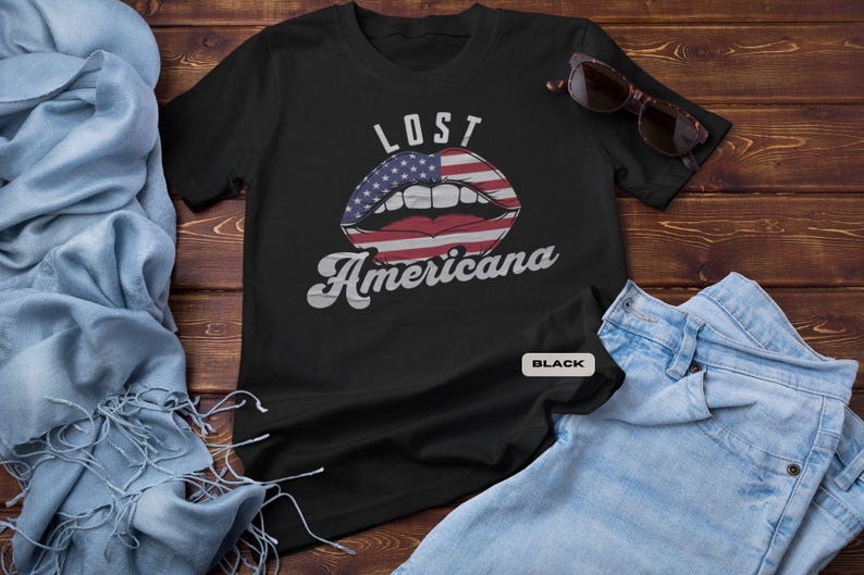 May include: Black t-shirt with the text "LOST Americana" above a graphic of lips designed with the American flag. The shirt is displayed on a wooden surface with sunglasses and denim jeans.