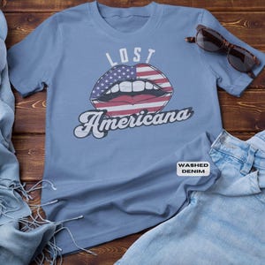 May include: A light blue t-shirt with the words "LOST Americana" and a graphic of lips with an American flag design. Sunglasses and denim jeans are next to the shirt.