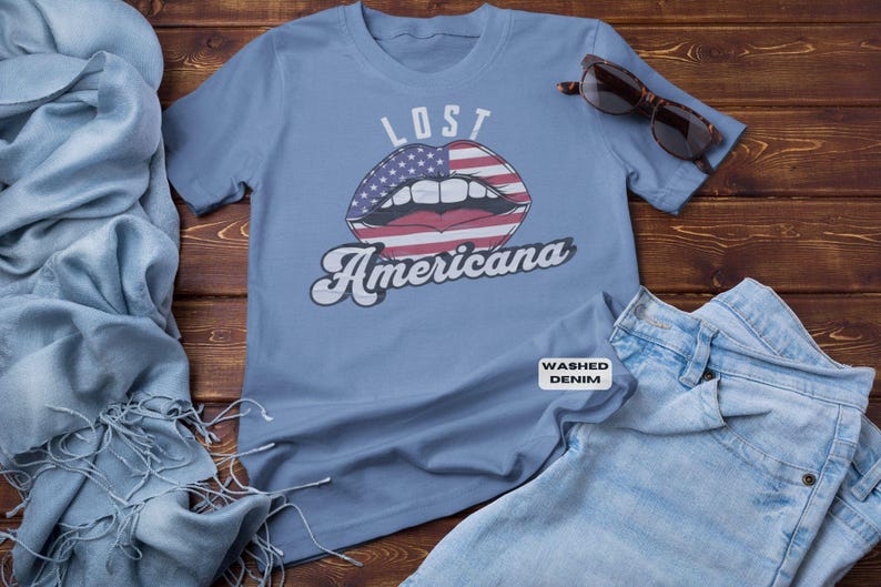 May include: A light blue t-shirt with the words "LOST Americana" and a graphic of lips with an American flag design. Sunglasses and denim jeans are next to the shirt.