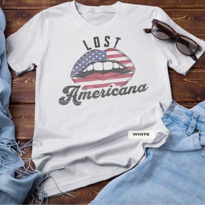 May include: White t-shirt with the text "LOST Americana" and a graphic of lips with an American flag design. The shirt is paired with blue jeans and sunglasses on a wooden surface.