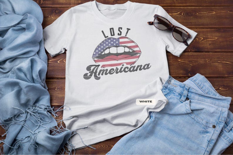 May include: White t-shirt with the text "LOST Americana" and a graphic of lips with an American flag design. The shirt is paired with blue jeans and sunglasses on a wooden surface.