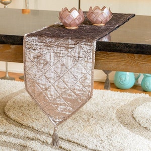 May include: A quilted, champagne-colored velvet table runner with a diamond pattern and a tassel. Two decorative lotus flower candle holders sit on the dark table. The runner is on a light-colored rug.