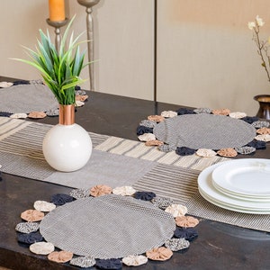 May include: Two black and white gingham placemats with a brown, black and white patterned fabric runner. The placemats have a decorative trim of beige, brown and black fabric rosettes. A white vase with a copper base holds a green plant.