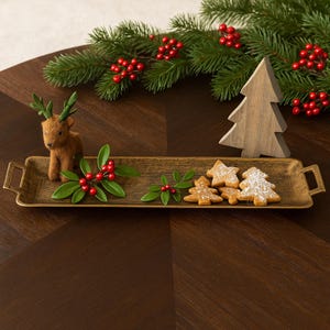 May include: A rectangular gold-toned serving tray with handles, decorated for the holidays. It holds a felt reindeer, holly sprigs, and tree-shaped cookies dusted with powdered sugar. A wooden tree and evergreen boughs with red berries complete the festive scene.