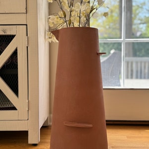 May include: A terracotta-colored ceramic vase with a wide mouth and a narrow base. The vase has a unique, slightly asymmetrical shape and a textured surface. It is filled with a bouquet of white flowers.