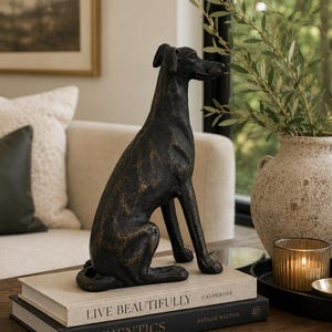 Peut inclure: Une figurine de chien texturée brun foncé en position assise, posée sur deux livres empilés intitulés Live Beautifully et Authentics. La sculpture présente une forme élancée et allongée avec une finition patinée, capturant le canidé de profil.