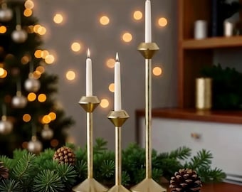 May include: Three gold candle holders with white candles. The candle holders are tall and slender with a geometric design. The candles are lit and the holders are arranged on a wooden table.