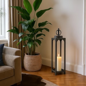 May include: A tall, black metal lantern with a lit white candle inside. The lantern has a decorative top and stands on a wooden floor. A large potted plant with green leaves is in the background.
