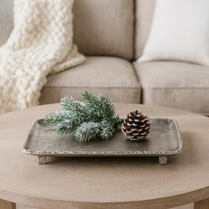 May include: A rectangular, silver-toned metal tray with a textured rim and small feet. The tray holds a sprig of frosted evergreen and a brown pinecone, suggesting a seasonal or holiday theme. The tray is on a light wood table.
