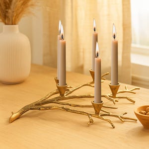 Peut inclure: Un chandelier doré en forme de branche supporte cinq bougies beiges allumées. Le chandelier est posé sur une surface en bois clair. Les bougies sont allumées d'une lueur chaleureuse, créant une ambiance chaleureuse. Décoration d'intérieur.