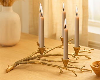 May include: A gold-colored, branch-shaped candelabra holds five lit, beige candles. The candelabra sits on a light-colored wooden surface. The candles are lit with a warm glow, creating a cozy ambiance. A decorative piece for home decor.