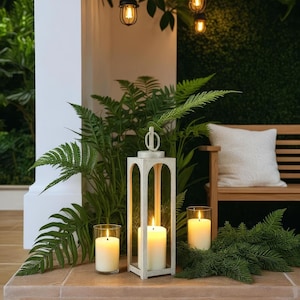 May include: A white wooden lantern with a lit candle inside, flanked by two glass candle holders with lit candles. Ferns and greenery surround the lantern, with a wooden bench and white pillow in the background. Overhead, there are decorative hanging lights.