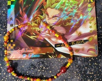 May include: A colorful beaded bracelet with red, orange, yellow, and brown beads. The bracelet has two larger red beads and a gold charm. The bracelet is lying on a black and white background with a card featuring an anime character in the background.