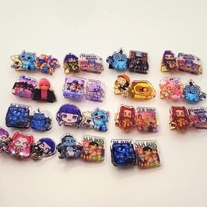May include: A collection of colorful acrylic pins featuring various cartoon characters and designs. The pins showcase a variety of colors and shapes, with some including text. The pins are arranged on a white surface.