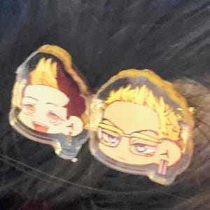 May include: Two cartoon character charms with yellow hair. The character on the left is wearing a black and white outfit. The character on the right is wearing glasses and holding a pair of chopsticks.