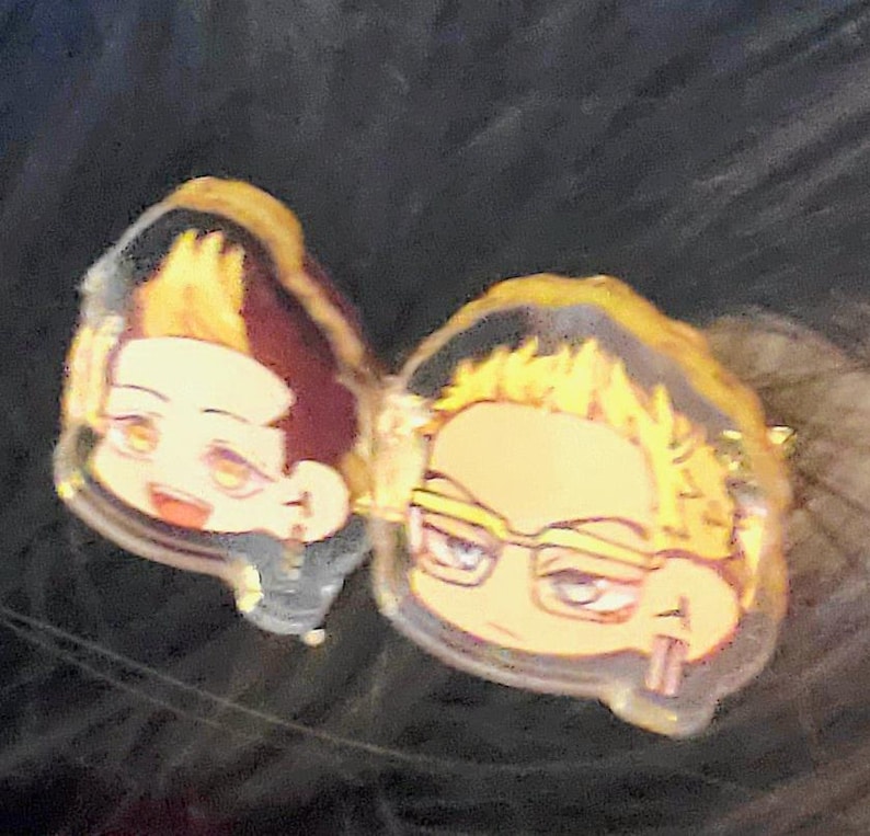 May include: Two cartoon character charms with yellow hair. The character on the left is wearing a black and white outfit. The character on the right is wearing glasses and holding a pair of chopsticks.