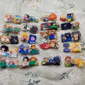 May include: A collection of anime-themed hair clips featuring various characters and scenes. The clips showcase vibrant colors and detailed artwork, with a variety of shapes and sizes. The clips are arranged on a floral patterned fabric.