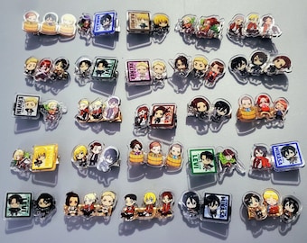 May include: A collection of clear acrylic pins featuring various anime characters. The pins showcase colorful illustrations of characters in different poses and settings. Some pins have text, such as character names. The pins are arranged on a light gray surface.