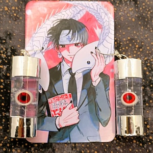 May include: Anime-themed earrings with a red eye design inside a clear, cylindrical container. A matching rectangular anime character image is in the background. The image features a character holding a mask and a book.