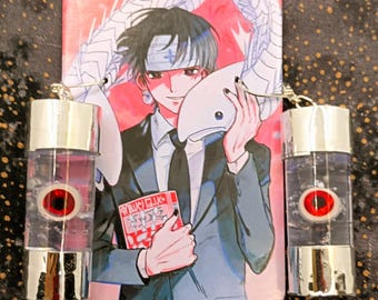 May include: Anime-themed earrings with a red eye design inside a clear, cylindrical container. A matching rectangular anime character image is in the background. The image features a character holding a mask and a book.