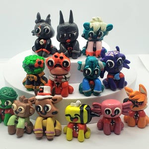 May include: A collection of 13 handmade polymer clay figurines. The figurines are all different colours and designs, including a green and red creature, a red and black bee, a pink axolotl, and a red and black fox.