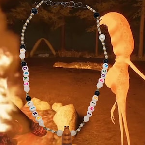 May include: A beaded necklace with the words "DON'T LIKE THIS ROCK" spelled out in colorful letter beads. The necklace features black and white beads, and a brown pendant. The background is a blurred outdoor scene.