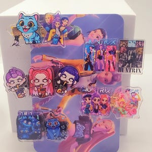May include: A collection of colorful acrylic charms featuring cartoon characters and the text "HUNTRIX". The charms include images of cats, people, and other designs. The background is a purple and blue image.