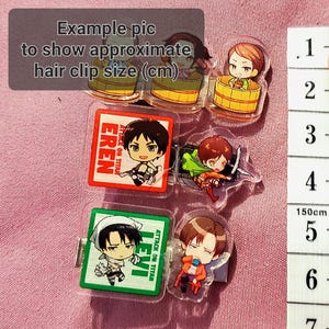May include: A set of six clear plastic hair clips featuring anime characters from the show "Attack on Titan". The clips are approximately 3 cm in size and feature the characters Eren, Levi, and a third character. The clips are displayed on a pink fabric background with a ruler for size reference.