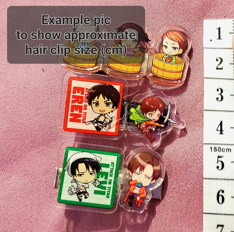 May include: A set of six clear plastic hair clips featuring anime characters from the show "Attack on Titan". The clips are approximately 3 cm in size and feature the characters Eren, Levi, and a third character. The clips are displayed on a pink fabric background with a ruler for size reference.