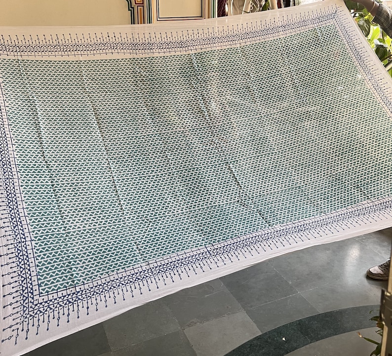 May include: White cotton fabric with a blue and green geometric pattern. The fabric has a border of small blue and white triangles.