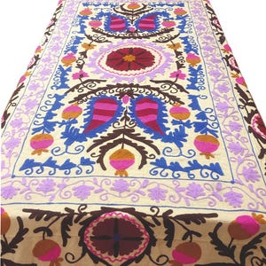 Può includere: Tessuto ricamato color crema con un motivo floreale centrale nei toni del rosso, rosa, blu e viola. L'intricato motivo comprende fiori e foglie stilizzate, creando una superficie vibrante e dettagliata.