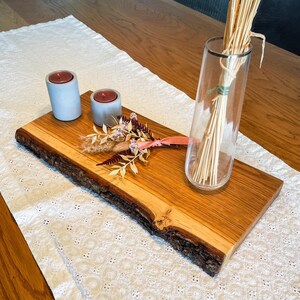 Könnte beinhalten: Ein Holzservierbrett mit rohem Rand, das zwei graue Betonkerzenhalter mit roten Kerzen, eine klare Glasvase mit getrockneten Stielen und dekorative Trockenblumen enthält. Das Brett liegt auf einem weißen Spitzentischläufer.
