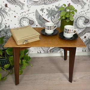 May include: A small, square wooden end table with rounded edges and dark brown legs. Two white coffee cups with black and brown geometric patterns are on black saucers on the table. A book titled "Craftsmanship in Wood" is on the table.