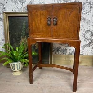 May include: A wooden cabinet with two doors featuring ornate dark metal drop handles, supported by a tall four-legged stand with a lower cross-stretcher. The wood has a warm brown finish with visible grain patterns throughout the structure.