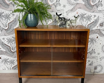 May include: A mid-century modern wooden cabinet with glass sliding doors. The cabinet has two shelves and sits on black legs. A green potted plant and small animal figurines are on top.