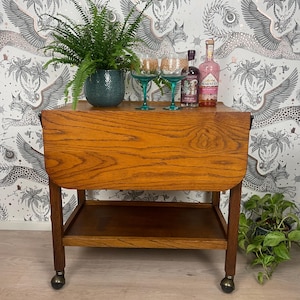 May include: A wooden bar cart with a drop-down top and a lower shelf. The cart has wheels and is made of dark wood with a natural finish. Two teal glasses and two bottles of pink liqueur are on the top shelf.