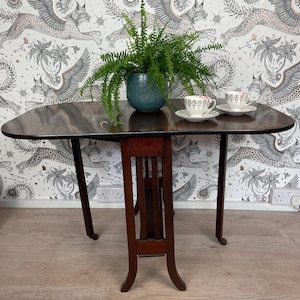 May include: A dark wood drop leaf table with a decorative carved centre support. The table has a rounded edge and sits on four legs with castors. Two teacups and saucers are on the table.