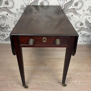 May include: A dark brown antique drop-leaf table with a single drawer. The table has a rectangular top and tapered legs with brass castors. The drawer features two round brass knobs and a keyhole. The table is made of wood.
