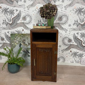 May include: A vintage-style, dark wood bedside cabinet with a single door and open shelf. The cabinet features carved detailing and brass fittings. A green vase with dried flowers, a book, and glasses are on top. A painting and a potted plant are nearby.