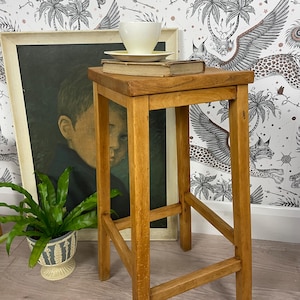 May include: A tall, four-legged wooden stool with a square seat. Resting on the seat is a thick, vintage hardback book topped with a plain, off-white ceramic teacup and saucer. The stool features a natural wood grain finish and simple joinery.